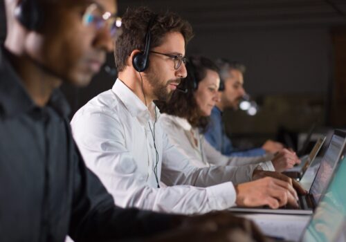 Side view of confident call center operator talking with client. Caucasian young man in eyeglasses typing on laptop while serving client. Call center concept
