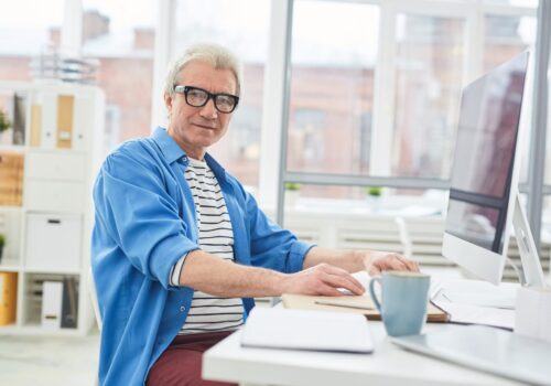 Confident mature boss in casualwear looking at camer aby workplace with computer monitor in front of him