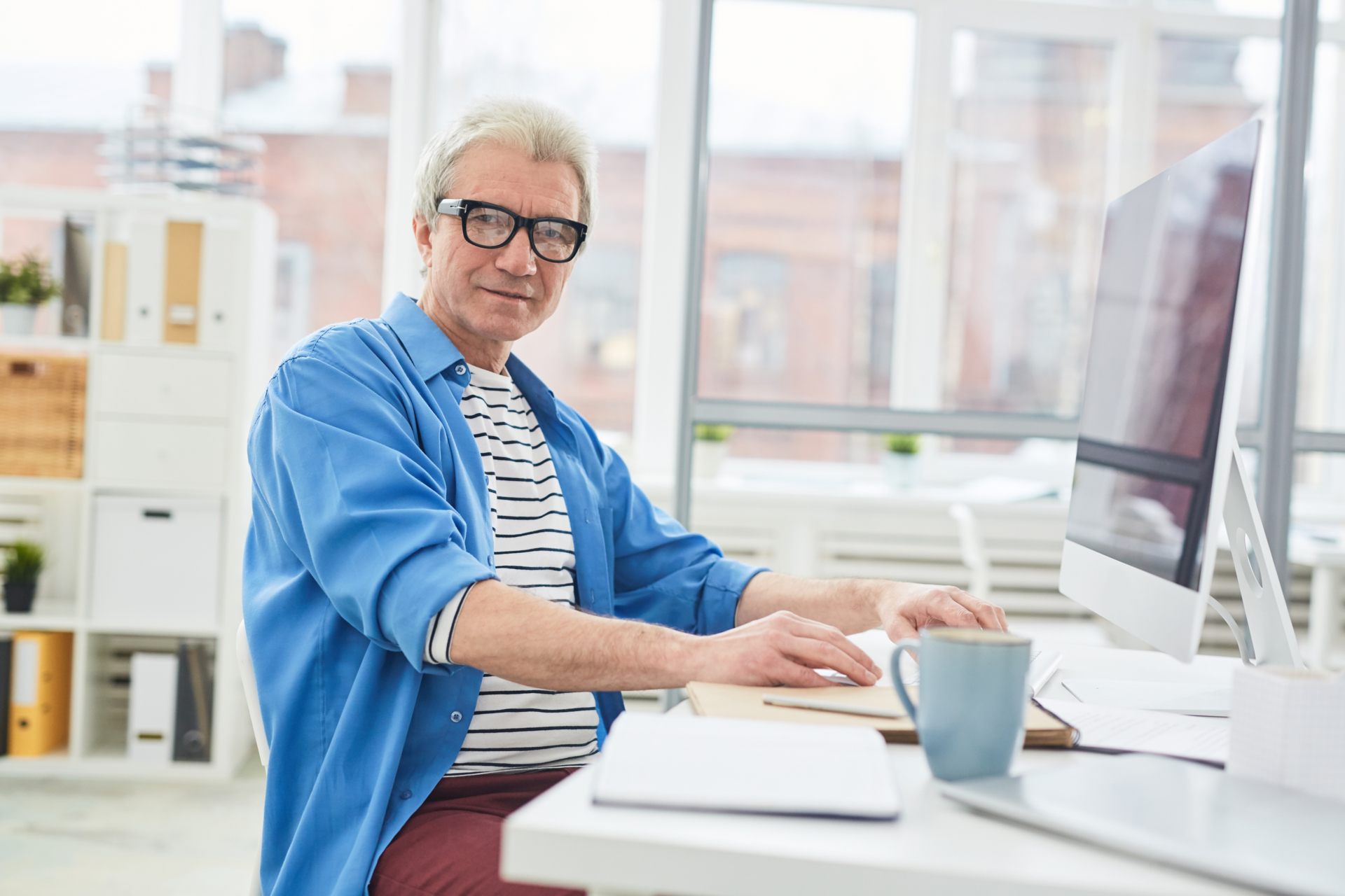 Confident mature boss in casualwear looking at camer aby workplace with computer monitor in front of him